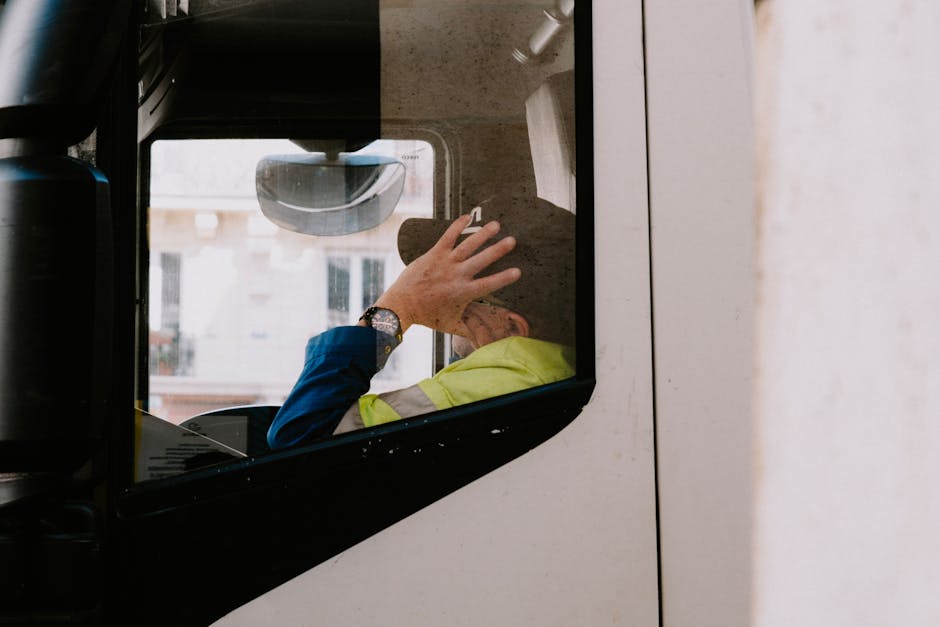 A truck driver in neon jacket rests with hand on head inside vehicle cabin, view from the side window.