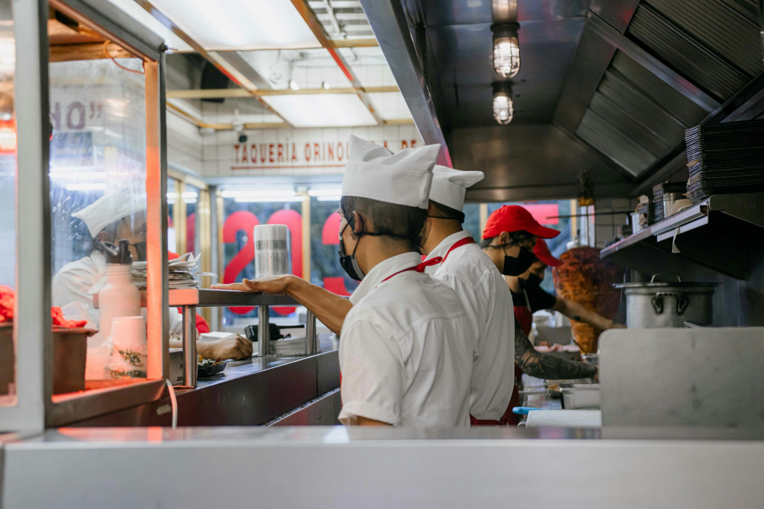 Culinary staff wearing uniforms and masks preparing meals in a bustling restaurant kitchen setting.