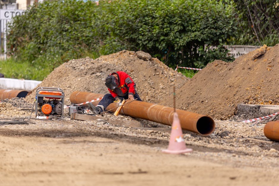 A construction worker welds a large pipe on a sunny day at an outdoor site.