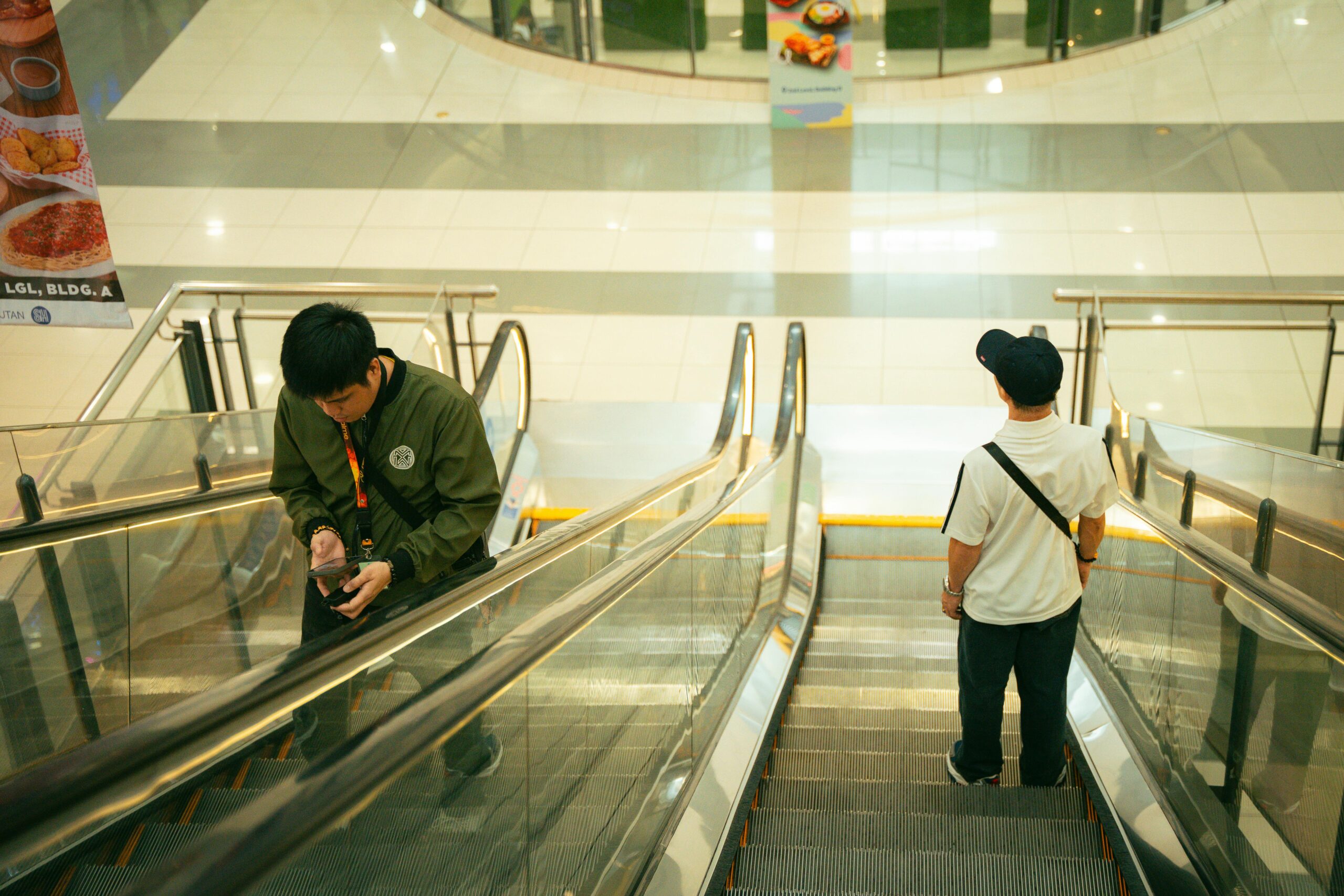Two men with smartphones on an escalator in a well-lit indoor shopping mall.