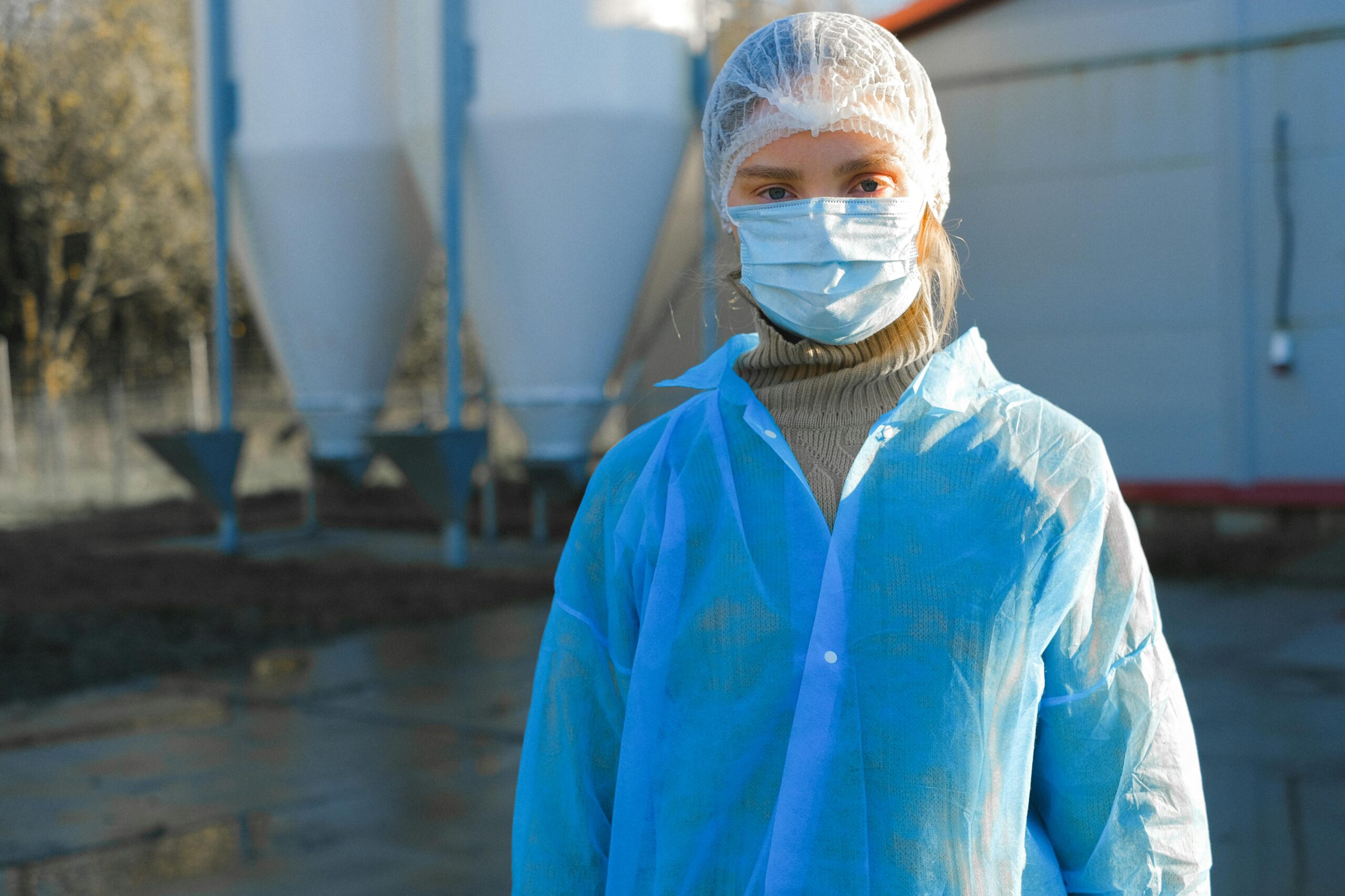 A woman wearing protective clothing, including a mask and hair net, outdoors at an industrial site.
